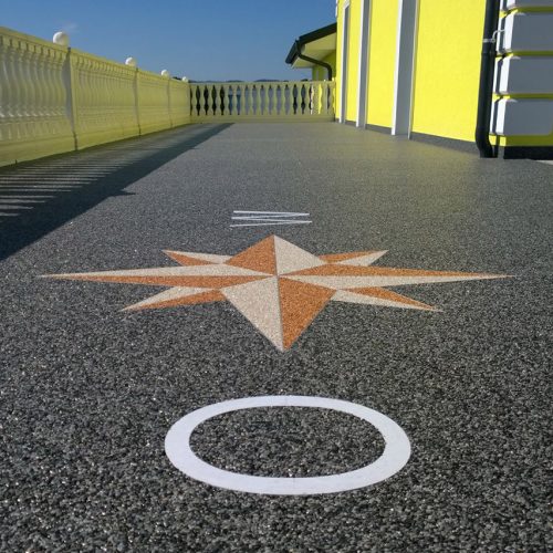 Der Natursteinteppich auf der gezeigten Terrasse ist anthrazitgrau und bildet einen strake Kontrast zur knallgelben Fassade und den weißen Balustraden. Einen eingearbeitete Windrose in beigen Farbtönen belebt das ganze Bild.