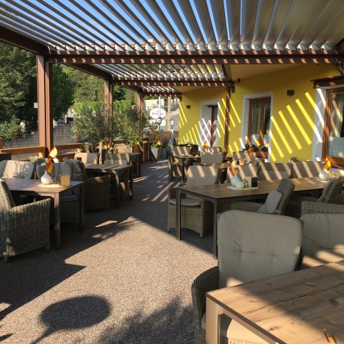 Das Bild zeigt einen Gastgarten mit Lamellenbeschattung. Der verlegte Steinteppich hat eine braun -graue Farbe