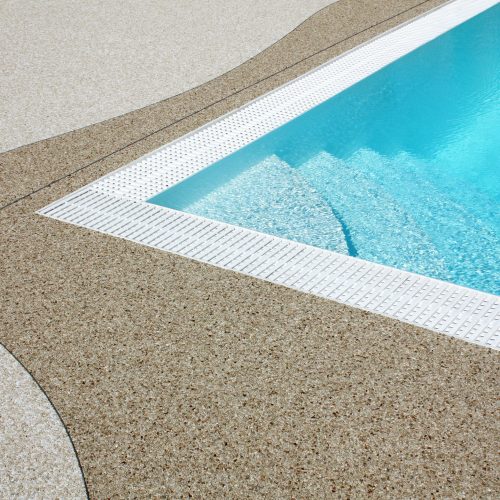 Ein dunkelbeiger Natursteinteppich umrandet mit geschwungener Linie ein Pool mit überlaufrinne. Der restliche Bereich wurde in einem hellern Farbeton verlegt