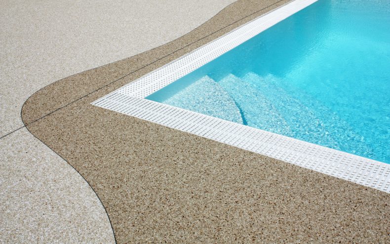 Ein dunkelbeiger Natursteinteppich umrandet mit geschwungener Linie ein Pool mit überlaufrinne. Der restliche Bereich wurde in einem hellern Farbeton verlegt